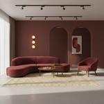 A modern living room scene featuring a curved upholstered sofa and a matching armchair in a muted terracotta red fabric, arranged on a patterned area rug with two small wooden coffee tables. The room has a terracotta-colored accent wall with arched recesses, a wall-mounted three-light sconce, and ceiling-mounted track lighting.