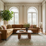 A styled living room featuring a large U-shaped sectional sofa upholstered in warm camel brown fabric, decorated with pink and patterned accent pillows. The sofa is positioned on a large patterned area rug in front of two tall arched windows with classical columns and a potted fiddle-leaf fig plant.
