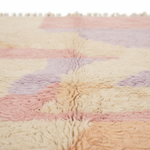 A close-up of a soft, textured area rug with a pastel geometric pattern in cream, pink, peach, and lavender, featuring a tassel fringe along the top edge.