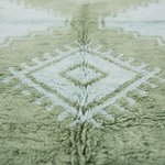 Close-up of a plush rug with a geometric diamond pattern in sage green and off-white, showing the soft texture of the pile.