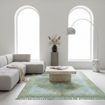 A modern living room scene featuring a light green and white patterned fringed area rug under a beige stone coffee table, next to a light gray sectional sofa and a tall silver floor lamp.