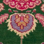 Close-up of a patterned rug with a green base, pink floral motifs, and a central purple and gold heart-shaped medallion.