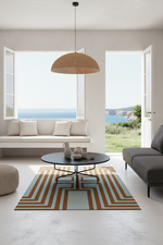 A modern living room interior with a geometric striped area rug in beige, brown, and light blue, featuring a round black coffee table, a white bench, and an open window overlooking the sea.
