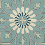 A close-up of a patterned rug featuring a central floral medallion in cream and navy blue, surrounded by a radial design in light blue, sage green, and taupe.