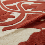 Close-up of a plush area rug with a raised floral or abstract scroll pattern in rust red and warm cream.