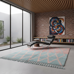A modern living room scene featuring a large shag area rug with a pink and light blue geometric pattern and fringe, styled with a black leather chaise lounge, a brick bookshelf, and an abstract painting.