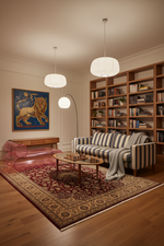 A cozy living room scene with a striped sofa, wooden bookshelves, a patterned area rug, pendant lights, and a glass coffee table. A pink translucent lounge chair sits to the left, and a framed lion painting hangs on the wall.