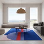 A modern living room with a large abstract area rug in blue, pink, and red tones, styled with a round coffee table, pendant light, and gray sofa. The room features large windows overlooking a coastal landscape.