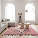 A pink geometric area rug with colorful diamond patterns and tassels is styled in a bright, modern living room with a light gray sectional sofa, a low wooden coffee table, and a tall silver floor lamp.