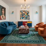 A styled living room interior with a curved teal sofa, two orange armchairs, a marble-top coffee table, a large green patterned area rug, wall sconces, and colorful abstract wall art in a white paneled room.