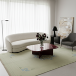 A modern living room scene featuring a large light sage green area rug with a geometric medallion pattern, styled with a cream curved sofa, a glossy dark red coffee table, a gray armchair, and a black floor lamp.