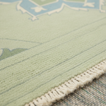 Close-up of a pale sage green area rug with a cream fringe edge and a subtle geometric pattern, layered over a textured gray rug underneath.