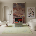 A styled living room featuring a large pale sage green area rug with a subtle blue and gold medallion pattern. The rug is styled in a modern living space with a stone coffee table, a red flower arrangement, a textured brown and gold marble fireplace wall, a white armchair, and a curved white sofa.