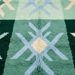 A close-up of a textured, loop-pile rug featuring a geometric pattern with sage green, forest green, light blue, and cream sections.