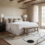 A styled bedroom interior featuring a bed dressed in white bedding, set with neutral beige and taupe pillows, a rustic wooden bench at the foot of the bed, a large abstract cream rug with black lines, and warm beige walls with exposed wood beams.