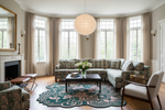 A beautifully styled living room featuring a large sectional sofa with a green and beige floral pattern, a matching accent chair, a black coffee table, and a decorative teal and pink rug. The room is illuminated by natural light from tall windows with beige curtains, and a large white paper lantern hangs from the ceiling.