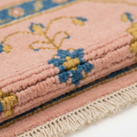 Close-up of a folded pink woven rug with cream fringe, blue and mustard floral motifs, and a patterned border.
