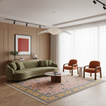 A styled living room interior featuring a curved olive green sofa, two orange armchairs, a patterned pink and blue area rug, a round wooden coffee table, and a wood-paneled wall with framed wall art.