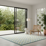 A large rectangular area rug with a geometric blue, green, and cream pattern is displayed in a modern room with large glass sliding doors overlooking a lush green garden. The room also features a wooden cane chair, a white side table, a floor lamp, and a white decorative screen.