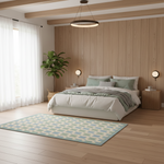 A modern bedroom scene with a bed dressed in white bedding and sage green pillows, set against a light wood-paneled wall. A light blue and cream geometric patterned area rug lies on the wood floor in front of the bed, and a tall potted plant stands near the window with sheer white curtains.