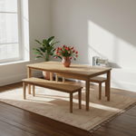 A light natural wood dining table with matching bench and chair, styled in a bright room with a window, potted plant, flowers, and books on a patterned rug.