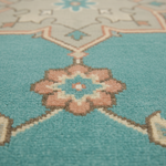 Close-up of a patterned area rug with a turquoise base and a central floral motif in dusty pink, cream, and brown.