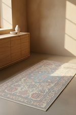 A rectangular area rug with a traditional floral pattern in muted blue, pink, and beige tones, styled on a neutral floor next to a wooden sideboard in a sunlit room.