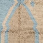 A close-up of a textured, loop-pile rug featuring a central beige chevron pattern flanked by light blue and cream zigzag stripes.