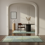 A styled interior scene showing a light blue and green patterned shag rug on a dark wood floor in the foreground, with a matching rug under a black round table and brown chair in the background. The room has cream walls with molding, a framed botanical print, and a white vase on a wooden pedestal.