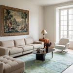 A styled living room featuring a large beige tufted sofa, a matching ottoman, a black oval coffee table, a light blue-green shag rug, a white modern armchair, and a large framed classical tapestry on the wall.
