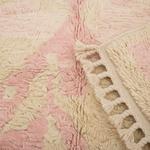 A close-up of a textured area rug with a soft, shaggy pile in alternating dusty pink and cream sections, finished with a cream-colored knitted border and tassel fringe.
