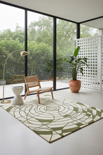 A modern area rug with an abstract green and cream geometric pattern, styled in a minimalist room with a wooden chair, floor lamp, and large windows overlooking greenery.
