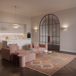 A modern living room featuring a dusty pink tufted sectional sofa and matching armchair on a large patterned oriental area rug. The room has dark wood herringbone floors, a minimalist white kitchen in the background, and a distinctive arched glass door.