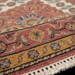 Close-up of a traditional patterned rug with cream, dusty pink, ochre yellow, and blue floral motifs and tasseled fringe.