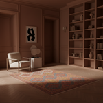 A styled interior room with warm brown walls, built-in wooden bookshelves, a cream armchair, a small side table with round lamps, and a large patterned area rug on a wood floor.