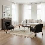 A styled living room interior featuring a cream sofa, two black armchairs with dark wood frames, a round coffee table, a patterned rug, and a fireplace with a large mirror, set against white walls and tall windows with sheer curtains.