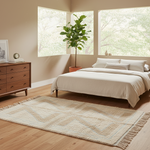 A styled bedroom scene with a bed dressed in white bedding, a cream woven area rug with fringe on a light wood floor, a wooden dresser, and a potted green plant near large windows.