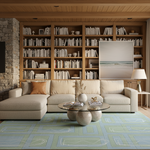 A staged living room interior featuring a large beige sectional sofa with a glass-top coffee table in front, set on a light blue and green geometric rug. Behind the sofa is a tall wooden bookcase filled with books, alongside a stone wall and a framed beach print. No people or animals are present.