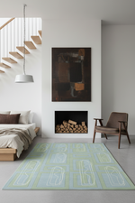 A modern area rug with a light blue and sage green geometric pattern is styled in a minimalist bedroom setting with a bed, staircase, fireplace, and armchair.