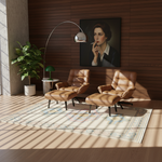 Two tan leather mid-century modern lounge chairs with matching ottomans are arranged on a patterned area rug in a sunlit room with wood-paneled walls. A large framed portrait hangs above, alongside a curved floor lamp, small side table with green lamp, and potted plant.