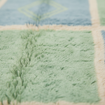 Close-up of a soft plush rug with a patchwork design, featuring a light green shaggy section, a dusty blue section with a faint diamond pattern, and cream-colored borders.