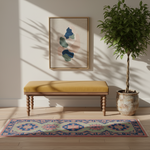 A styled interior scene with a wooden bench featuring a mustard yellow cushion, a colorful patterned rug, a potted tree, and framed abstract wall art against a warm beige wall with leaf shadows.