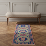 A long rectangular area rug with a geometric and floral pattern in sage green, navy, pink, and blue, styled on a light wood herringbone floor in front of a beige upholstered bench.