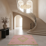 A pink rectangular area rug with a green geometric diamond pattern and fringe, displayed on a light stone floor in a grand, elegant staircase hall with beige walls and a black iron railing.