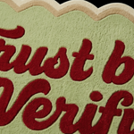 A close-up of a fuzzy, cloud-shaped sign or mat in pale sage green with raised red cursive lettering that reads 'Trust but Verifi'.