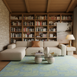 A modern living room featuring a large beige sectional sofa with a brown accent pillow, set against a wall of wooden bookshelves. A light blue and green patterned rug covers the floor, with a glass-top coffee table in front. A gold floor lamp and a framed beach print sit on a small side table.