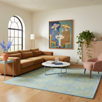 A styled living room featuring a large tan sectional sofa, a light blue and green patterned area rug, a pink armchair, and a framed colorful painting on the wall.
