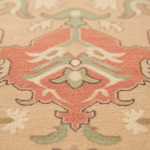 Close-up of a patterned area rug with a central coral-red medallion, sage green accents, and cream floral details on a warm beige background.