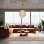 A modern sectional sofa in a warm rust-brown color arranged in an L-shape in a contemporary loft living room with large windows, a concrete ceiling, a hanging paper lantern pendant light, and a patterned area rug.