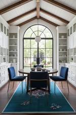 A dining room with a round wooden table and three blue upholstered chairs arranged on a teal floral area rug. The room has white cabinetry, a large arched window, and a high wood-beamed ceiling.